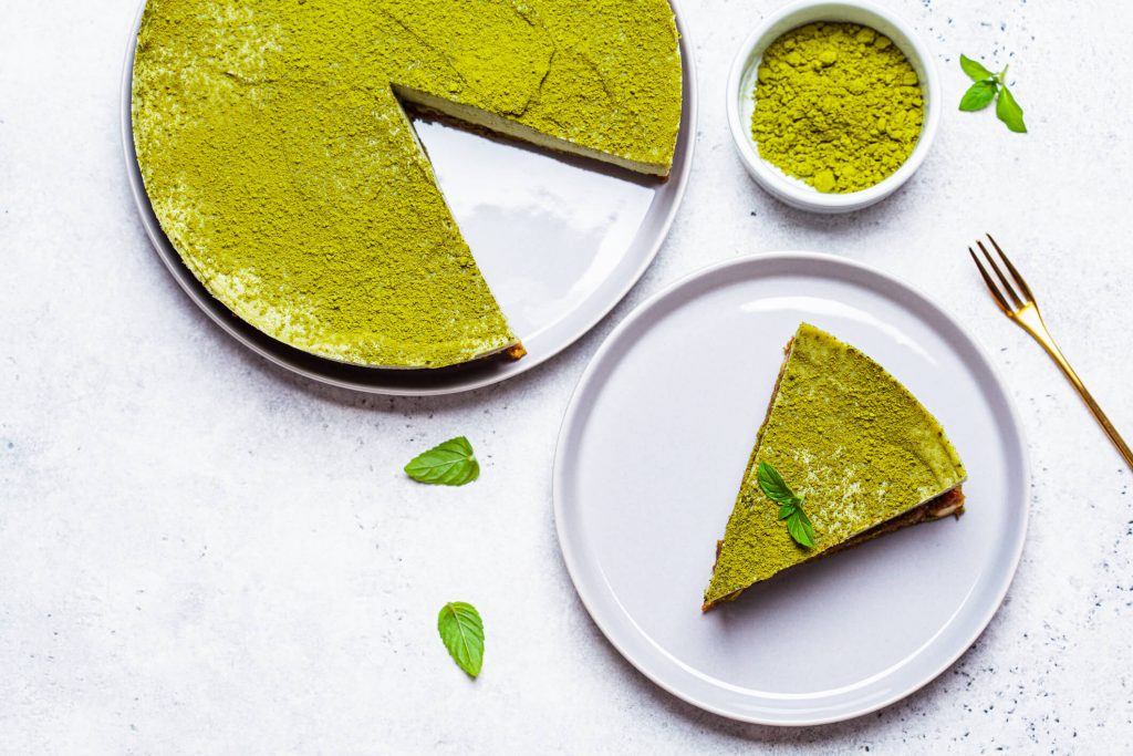 A cheesecake topped with matcha garnished with green leaves, with a bowl of matcha powder beside it. One slice is taken out and set on a plate. 