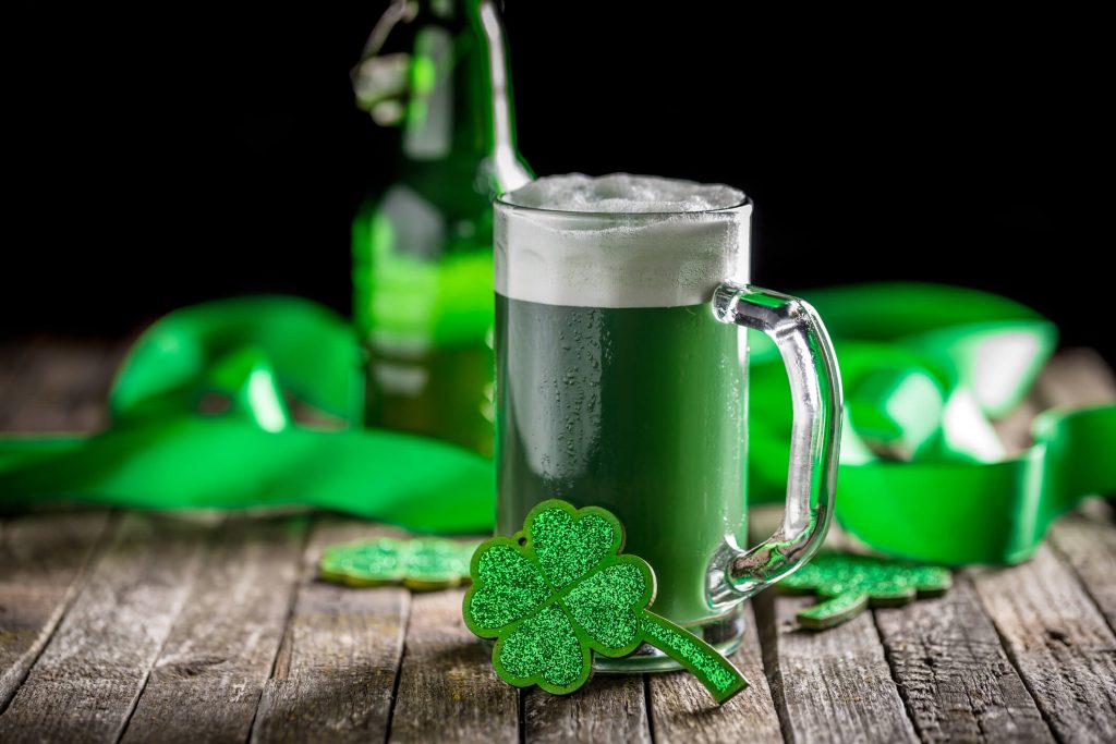 A glass of green beer with a shamrock decoration on it, various green Saint Patrick's Day themed items in the background.