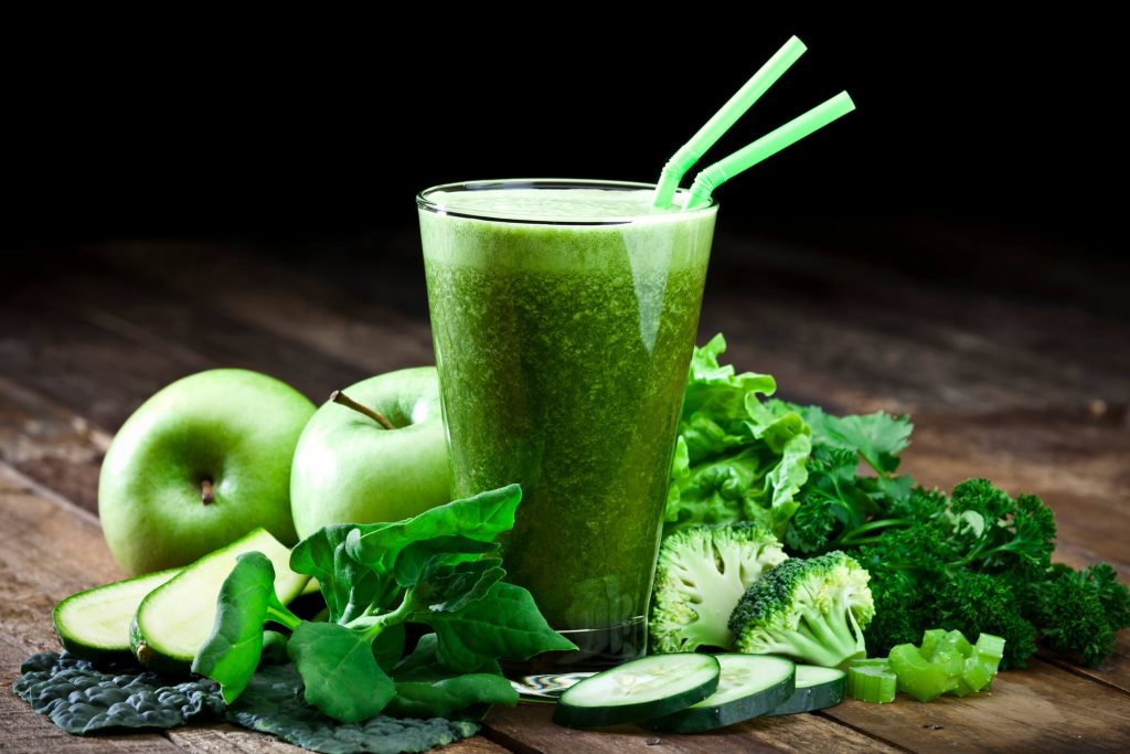 A green smoothie with 2 green straws and various green ingredients in the background