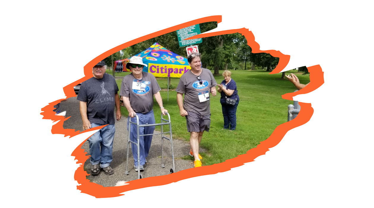 Three participants including Paul Fireman smile as they walk in the .5K. They are in a park scene with a portable tent and spectators in the background.