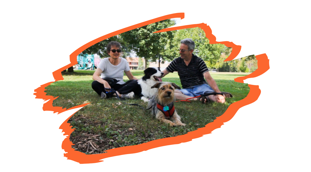 Two spectators sit in a grassy, shady area of Westinghouse park with their two dogs. The spectator on the left is wearing sunglasses and sitting cross-legged. The spectator on the right is wearing prescription glasses with convertible lenses, sitting and petting one of the dogs.