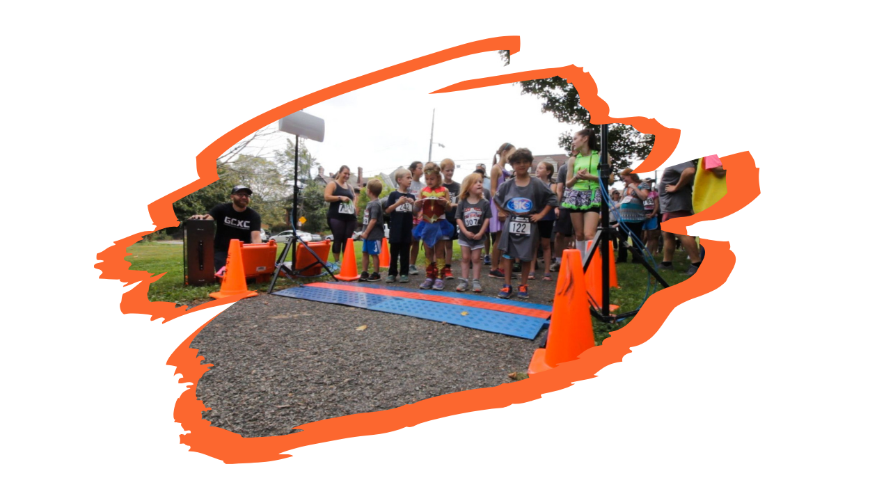 Young participants wait at the What's the Point .5K starting line. There are adult participants at the sides and in the background supervising. The starting line is flanked by orange traffic cones and tripods.